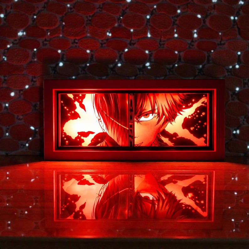 Pu&ograve; includere: Una scatola luminosa in stile anime con cornice rossa con un'immagine divisa del volto di un personaggio, nei toni del rosso e dell'arancione. L'immagine si riflette su una superficie lucida sottostante. Lo sfondo &egrave; un muro rosso strutturato con piccole luci bianche.
