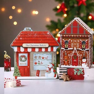 May include: Two Christmas-themed tins in the shape of buildings. One is a red bakery with the text "The Christmas Bakery" and a snowman. The other is a gingerbread house with festive details.