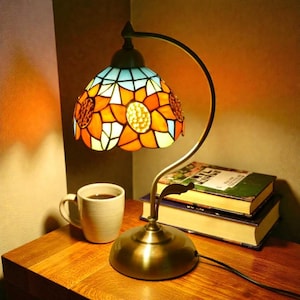 Può includere: Una lampada da tavolo in vetro colorato con un motivo a girasole. La lampada ha una base in bronzo e un braccio curvo. Il paralume presenta pezzi di vetro arancione, giallo, bianco e blu. Una tazza bianca di caffè e due libri sono sul tavolo di legno.
