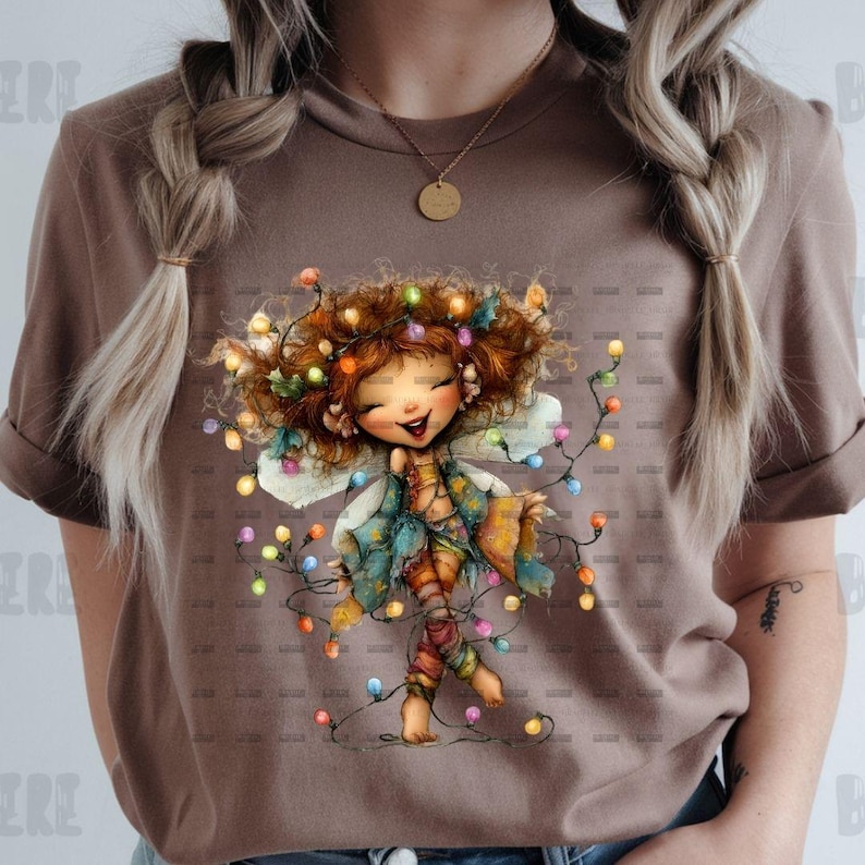 May include: A brown t-shirt featuring a whimsical illustration of a fairy adorned with colorful lights. The fairy has red hair, a smiling face, and is wearing a blue and orange outfit. The fairy is holding a string of lights.