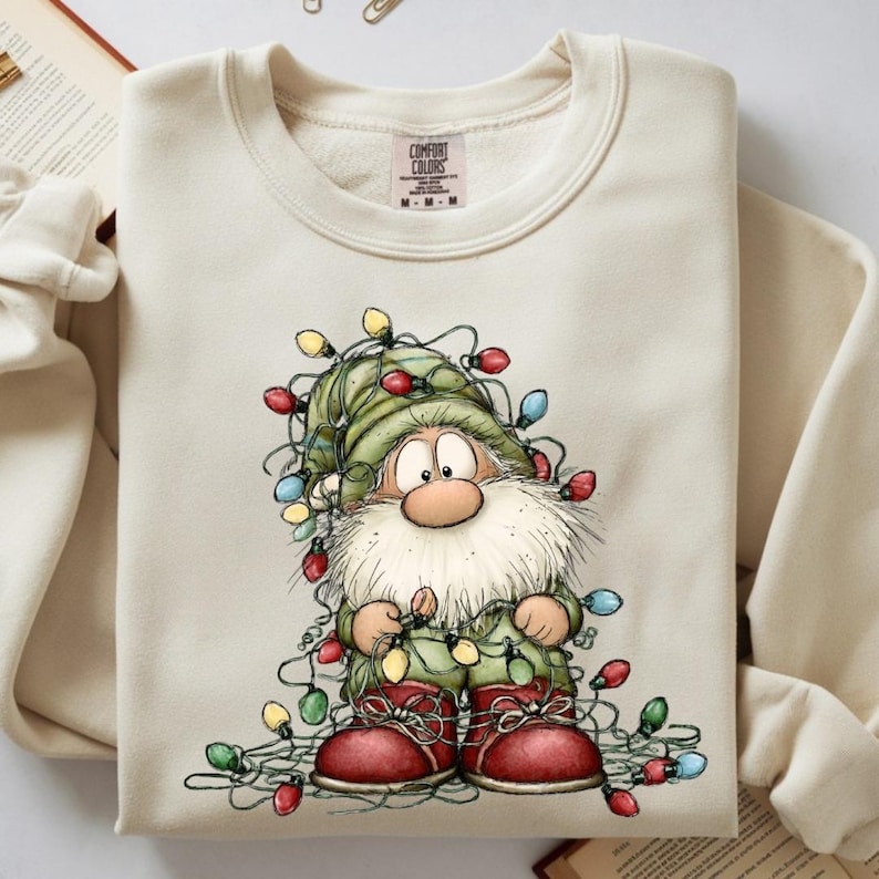 May include: Cream-coloured sweatshirt with a cartoon gnome entangled in colourful Christmas lights. The gnome wears a green hat, matching outfit, and red boots. The Comfort Colors brand tag is visible at the top.