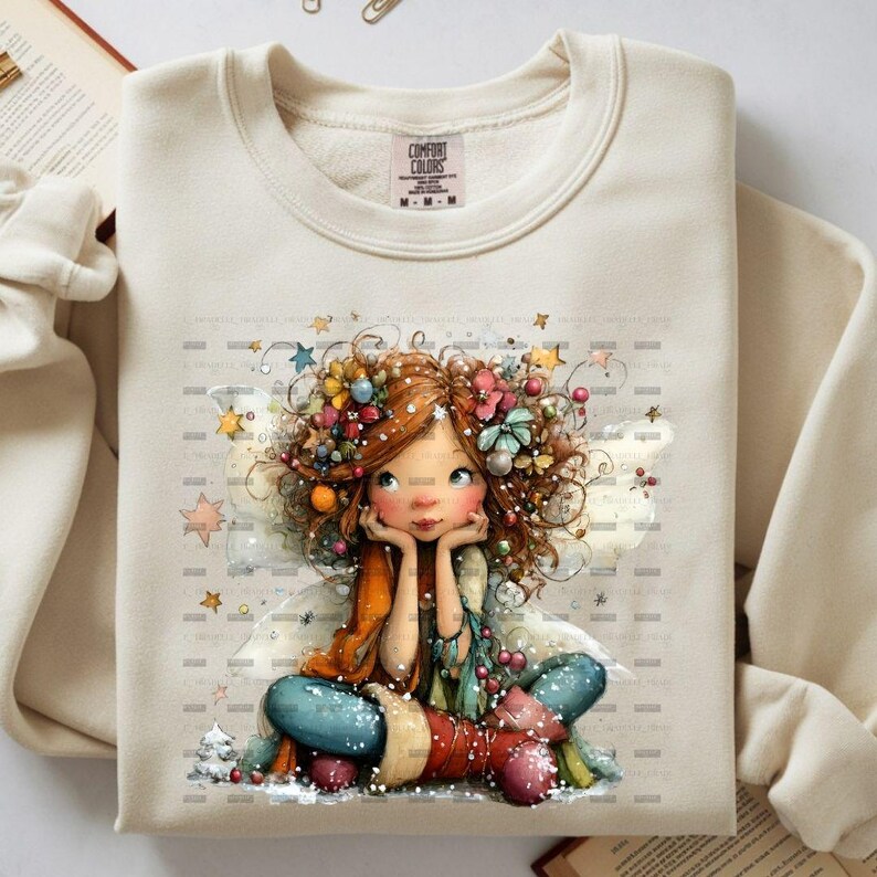 May include: Cream-colored Comfort Colors sweatshirt featuring a whimsical illustration of a fairy with red hair adorned with flowers and stars. The fairy is seated, resting her chin on her hands, with a colorful outfit and wings. The sweatshirt has a crew neck.