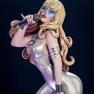 Puede incluir: Estatua de una artista con un mono plateado, sosteniendo un micrófono. La figura tiene cabello rubio, pintura facial azul y joyas plateadas. La estatua está posada en una postura dinámica sobre un fondo oscuro.