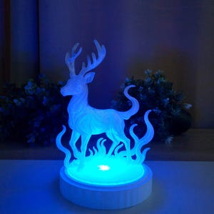 Könnte beinhalten: Eine leuchtend blaue Hirschfigur mit filigranen Details, die auf einem weißen Sockel steht. Der Hirsch wird von blauem Licht beleuchtet, und der Sockel hat einen blauen Lichtring. Der Gesamteffekt ist ein magischer, ätherischer Schein.