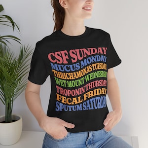 Op de afbeelding: Zwart t-shirt met kleurrijke tekst. De tekst bevat medische termen voor elke dag van de week: CSF zondag, Mucus maandag, Trichomonas dinsdag, Wet Mount woensdag, Troponine donderdag, Fecal vrijdag en Sputum zaterdag.