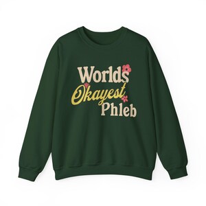 Puede incluir: Una sudadera verde oscuro con el texto "World's Okayest Phleb" en letras crema y amarillas, acentuado con gráficos florales rosas. La sudadera es de cuello redondo.