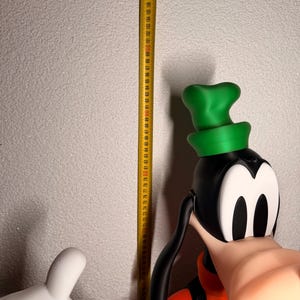 Puede incluir: Una figura tridimensional grande de Goofy, un personaje de dibujos animados, con cabeza negra, sombrero verde y atuendo naranja. Una cinta m&eacute;trica amarilla se coloca junto a la figura, mostrando medidas en cent&iacute;metros.