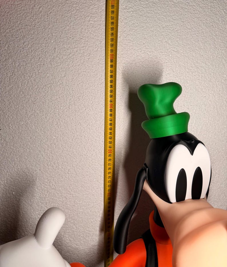 Puede incluir: Una figura tridimensional grande de Goofy, un personaje de dibujos animados, con cabeza negra, sombrero verde y atuendo naranja. Una cinta m&eacute;trica amarilla se coloca junto a la figura, mostrando medidas en cent&iacute;metros.