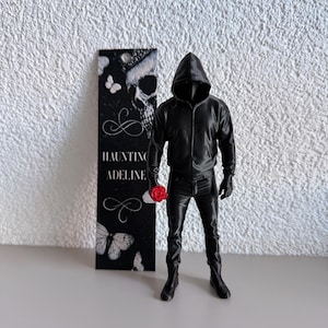 Peut inclure: Une figurine noire portant une veste à capuche, un pantalon et des bottes, tenant une petite rose rouge. À côté se trouve un marque-page noir rectangulaire orné d'un crâne blanc, de papillons et du texte Haunting Adeline en blanc.
