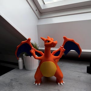 Könnte beinhalten: Eine orangefarbene Charizard-Figur mit blauen Flügelakzenten und einem gelben Bauch. Die Figur ist mit erhobenen Armen posiert und zeigt ihre Flügel. Die Figur steht auf einer grauen Oberfläche.