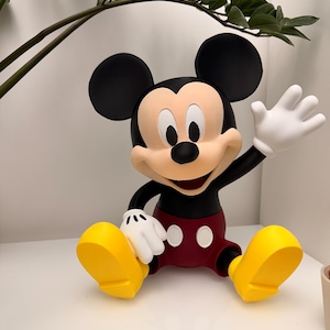 Op de afbeelding: Een Mickey Mouse-beeldje in zittende positie, zwaaiend met &eacute;&eacute;n hand. De figuur heeft zwarte oren, een zwarte torso, een rode korte broek, gele schoenen en witte handschoenen. Het beeldje staat op een wit oppervlak.