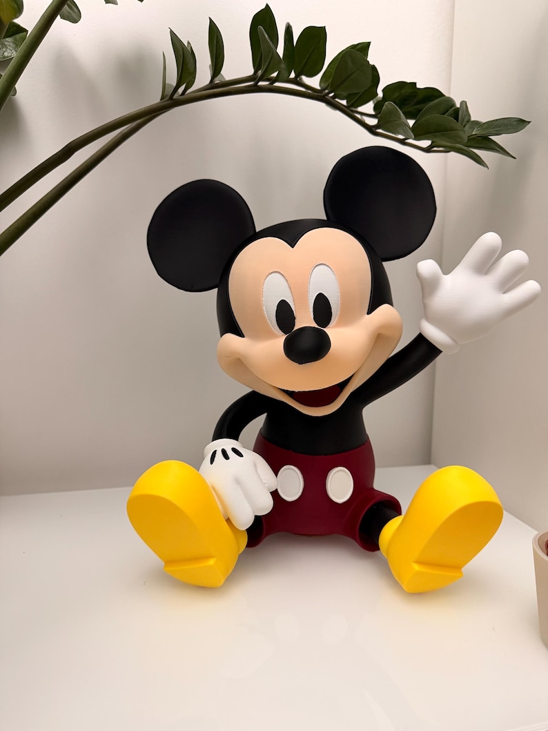 Op de afbeelding: Een Mickey Mouse-beeldje in zittende positie, zwaaiend met &eacute;&eacute;n hand. De figuur heeft zwarte oren, een zwarte torso, een rode korte broek, gele schoenen en witte handschoenen. Het beeldje staat op een wit oppervlak.