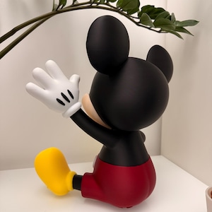 Op de afbeelding: Een zwarte Mickey Mouse-beeldje met witte handschoenen, een rode broek en gele schoenen. Het beeldje poseert met &eacute;&eacute;n arm omhoog, alsof het zwaait. Het beeldje is een verzamelobject.