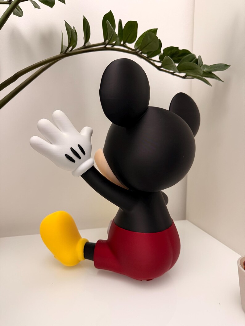 Op de afbeelding: Een zwarte Mickey Mouse-beeldje met witte handschoenen, een rode broek en gele schoenen. Het beeldje poseert met &eacute;&eacute;n arm omhoog, alsof het zwaait. Het beeldje is een verzamelobject.