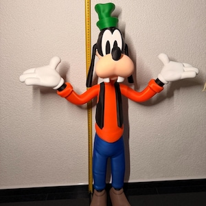 Puede incluir: Una gran figura de Goofy, con un sombrero verde, orejas negras y una gran nariz naranja. Viste una camisa naranja, chaleco negro, pantalones azules y zapatos marrones. Sus manos enguantadas blancas est&aacute;n extendidas. Una cinta m&eacute;trica amarilla es visible en el fondo.