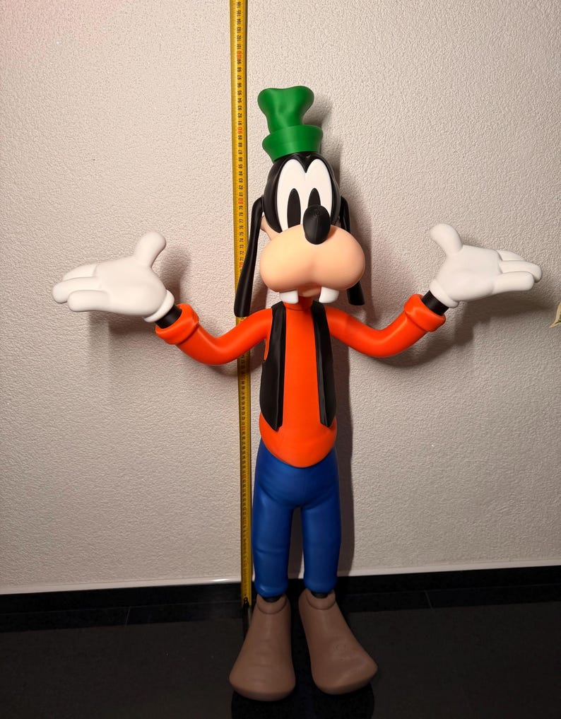Puede incluir: Una gran figura de Goofy, con un sombrero verde, orejas negras y una gran nariz naranja. Viste una camisa naranja, chaleco negro, pantalones azules y zapatos marrones. Sus manos enguantadas blancas est&aacute;n extendidas. Una cinta m&eacute;trica amarilla es visible en el fondo.