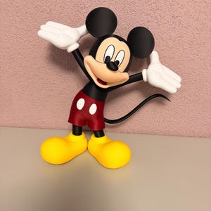 Puede incluir: Una figura de Mickey Mouse en una pose de bienvenida. La figura tiene orejas negras, un cuerpo negro, pantalones cortos rojos con botones blancos, zapatos amarillos y guantes blancos. El fondo es una pared rosa texturizada.