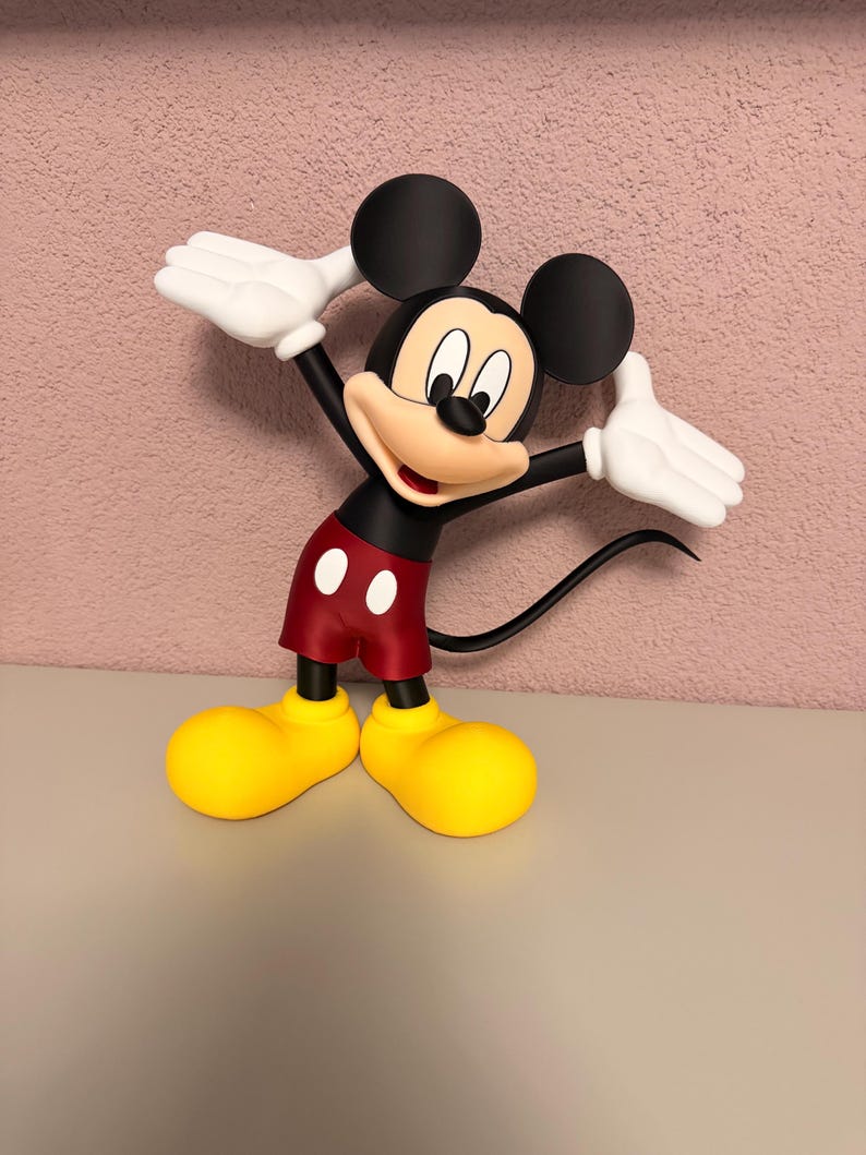Puede incluir: Una figura de Mickey Mouse en una pose de bienvenida. La figura tiene orejas negras, un cuerpo negro, pantalones cortos rojos con botones blancos, zapatos amarillos y guantes blancos. El fondo es una pared rosa texturizada.