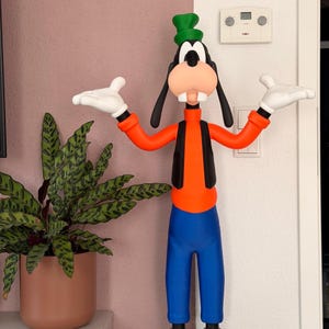 Puede incluir: Una colorida figura de Goofy, un personaje de dibujos animados de Disney, est&aacute; de pie con los brazos extendidos. La figura lleva un sombrero verde, una camisa naranja, un chaleco negro, pantalones azules y zapatos marrones. Guantes blancos y una gran cara sonriente completan el dise&ntilde;o.