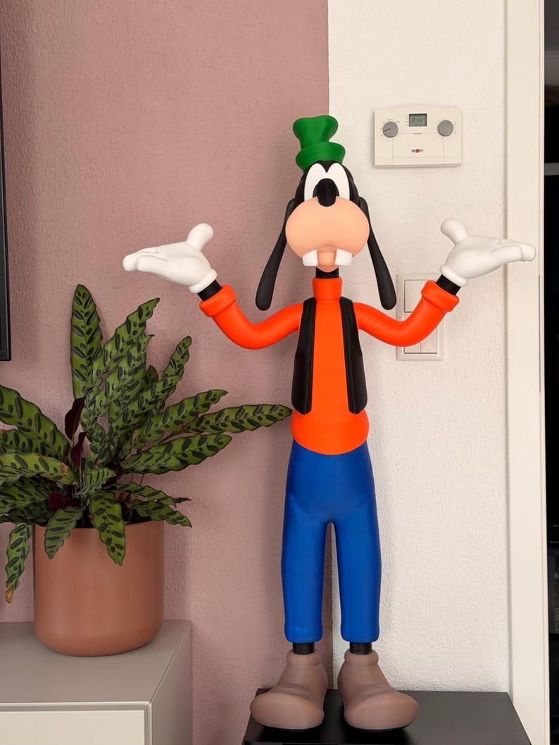 Puede incluir: Una colorida figura de Goofy, un personaje de dibujos animados de Disney, est&aacute; de pie con los brazos extendidos. La figura lleva un sombrero verde, una camisa naranja, un chaleco negro, pantalones azules y zapatos marrones. Guantes blancos y una gran cara sonriente completan el dise&ntilde;o.