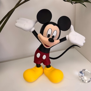 Puede incluir: Una figura de Mickey Mouse con los brazos levantados. La figura tiene una cabeza negra con orejas grandes, una camisa roja, pantalones cortos rojos con botones blancos, zapatos amarillos y guantes blancos. Sobre una superficie blanca.