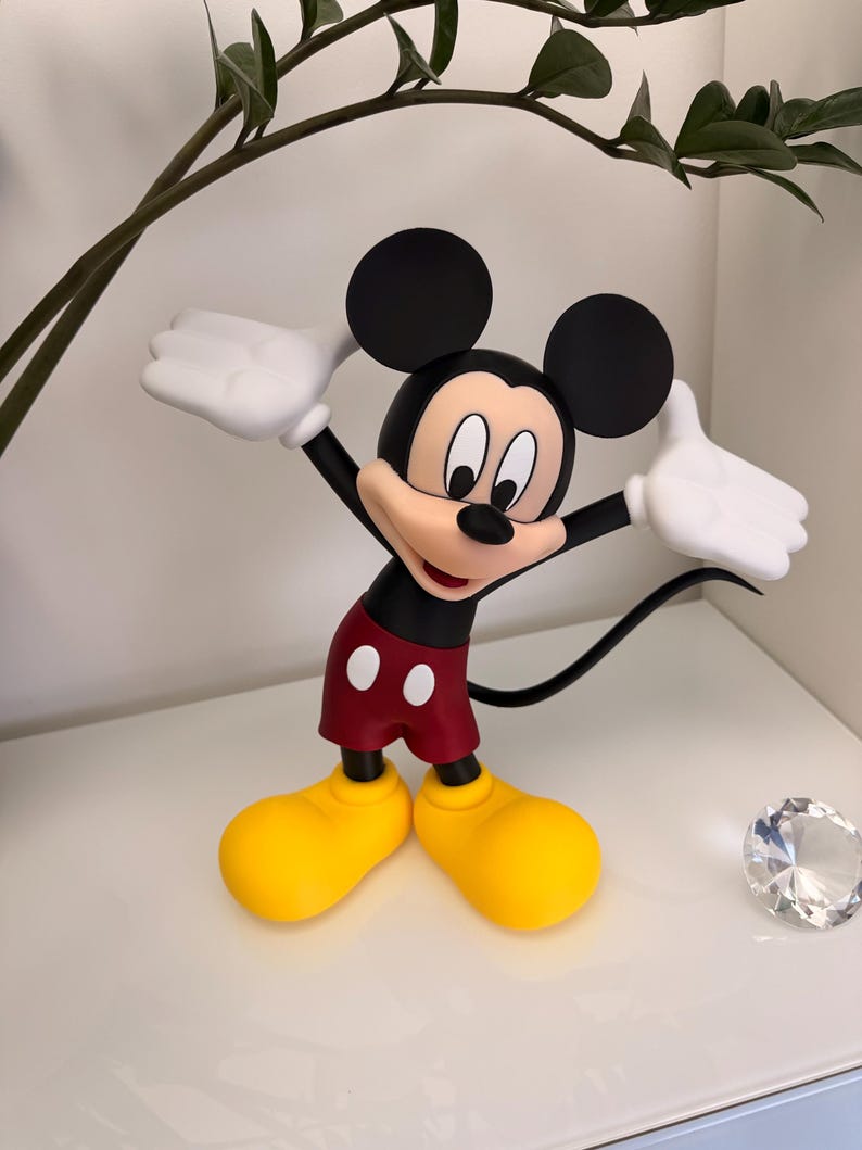 Puede incluir: Una figura de Mickey Mouse con los brazos levantados. La figura tiene una cabeza negra con orejas grandes, una camisa roja, pantalones cortos rojos con botones blancos, zapatos amarillos y guantes blancos. Sobre una superficie blanca.