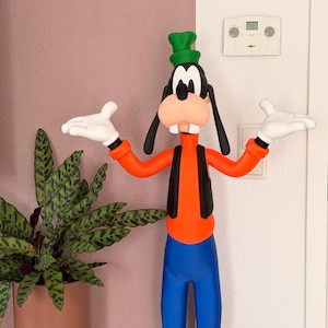 Puede incluir: Una colorida figura de Goofy, un personaje de dibujos animados de Disney, de pie con los brazos extendidos. Viste una camisa naranja, chaleco negro, pantalones azules y un sombrero verde. La figura est&aacute; junto a una planta en maceta.