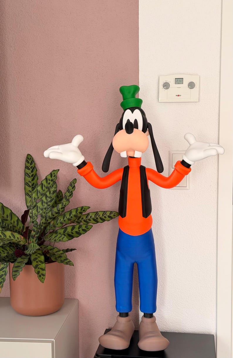 Puede incluir: Una colorida figura de Goofy, un personaje de dibujos animados de Disney, de pie con los brazos extendidos. Viste una camisa naranja, chaleco negro, pantalones azules y un sombrero verde. La figura est&aacute; junto a una planta en maceta.