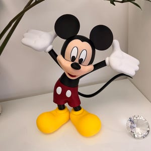 Puede incluir: Una figura de Mickey Mouse con los brazos extendidos, vistiendo una camiseta negra, pantalones cortos rojos con botones blancos, zapatos amarillos y guantes blancos. El ic&oacute;nico personaje tiene grandes orejas negras y una expresi&oacute;n sonriente. Un diamante decorativo est&aacute; al lado de la figura.