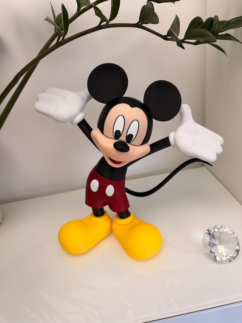 Puede incluir: Una figura de Mickey Mouse con los brazos extendidos, vistiendo una camiseta negra, pantalones cortos rojos con botones blancos, zapatos amarillos y guantes blancos. El ic&oacute;nico personaje tiene grandes orejas negras y una expresi&oacute;n sonriente. Un diamante decorativo est&aacute; al lado de la figura.