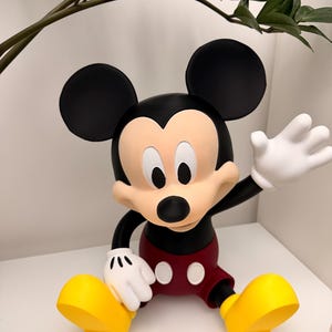 Op de afbeelding: Een Mickey Mouse-beeldje in een zittende pose. De figuur heeft een zwarte kop en oren, een bruin gezicht, witte handschoenen, een rode korte broek en gele schoenen. Het beeldje staat op een wit oppervlak.