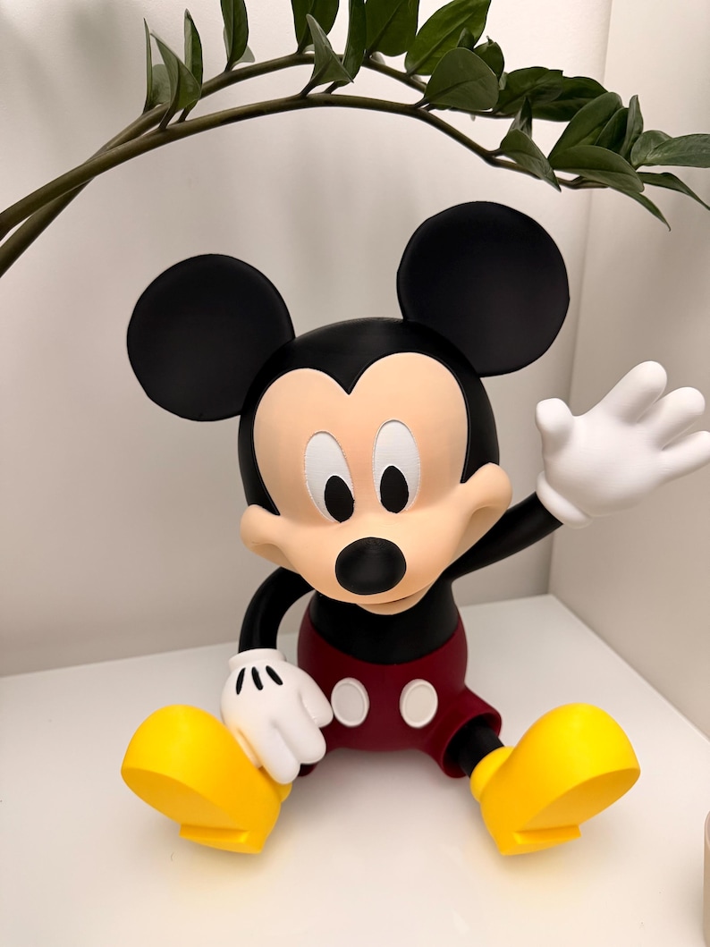 Op de afbeelding: Een Mickey Mouse-beeldje in een zittende pose. De figuur heeft een zwarte kop en oren, een bruin gezicht, witte handschoenen, een rode korte broek en gele schoenen. Het beeldje staat op een wit oppervlak.