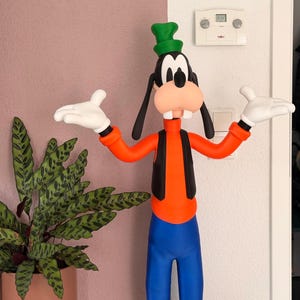 Puede incluir: Una colorida figura de Goofy, un personaje de dibujos animados de Disney, de pie con los brazos extendidos. Viste una camisa naranja, pantalones azules, un chaleco negro y un sombrero verde. Sus grandes rasgos de dibujos animados incluyen una gran nariz y guantes blancos. Una planta en maceta est&aacute; a la izquierda.