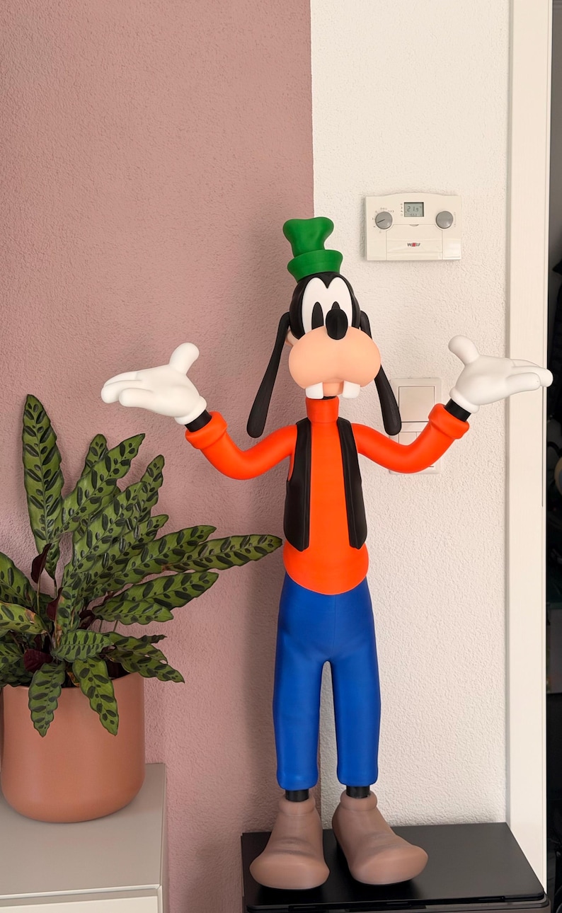 Puede incluir: Una colorida figura de Goofy, un personaje de dibujos animados de Disney, de pie con los brazos extendidos. Viste una camisa naranja, pantalones azules, un chaleco negro y un sombrero verde. Sus grandes rasgos de dibujos animados incluyen una gran nariz y guantes blancos. Una planta en maceta est&aacute; a la izquierda.