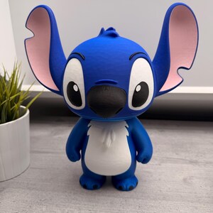 Peut inclure: Figurine bleue repr&eacute;sentant le personnage de Stitch, avec de grandes oreilles aux int&eacute;rieurs roses, de grands yeux et un nez noir. La figurine a un ventre blanc et se tient debout. Objet de collection.