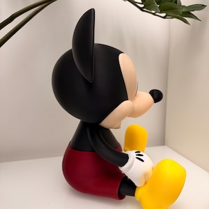 Op de afbeelding: Een zittend Mickey Mouse-beeldje. De figuur heeft een zwarte kop en oren, een beige gezicht en een zwarte neus. Het draagt een rode korte broek, zwarte armen, witte handschoenen en gele schoenen. Het beeldje staat op een wit oppervlak.