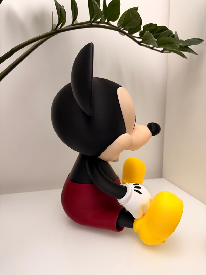 Op de afbeelding: Een zittend Mickey Mouse-beeldje. De figuur heeft een zwarte kop en oren, een beige gezicht en een zwarte neus. Het draagt een rode korte broek, zwarte armen, witte handschoenen en gele schoenen. Het beeldje staat op een wit oppervlak.