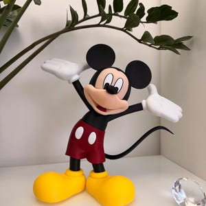 Puede incluir: Una figura de Mickey Mouse en una pose de bienvenida. La figura tiene orejas negras, pantalones cortos rojos, zapatos amarillos y guantes blancos. La figura est&aacute; sobre una superficie blanca con una planta en el fondo.