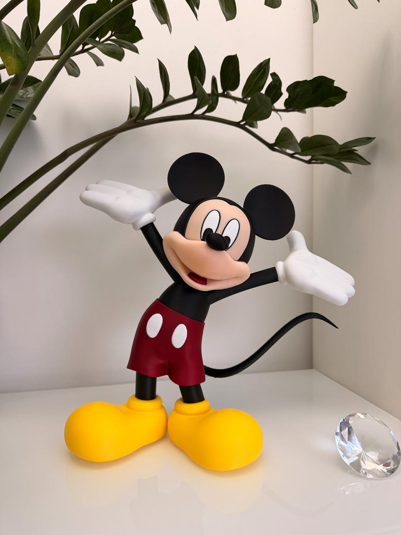 Puede incluir: Una figura de Mickey Mouse en una pose de bienvenida. La figura tiene orejas negras, pantalones cortos rojos, zapatos amarillos y guantes blancos. La figura est&aacute; sobre una superficie blanca con una planta en el fondo.