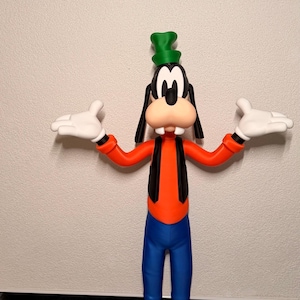 Puede incluir: Una figura de Goofy, un personaje de Disney, de pie con los brazos extendidos. La figura lleva un sombrero verde, orejas negras y un chaleco negro sobre una camisa naranja. Pantalones azules y zapatos marrones completan el look. La figura tiene guantes blancos y una gran sonrisa amistosa.