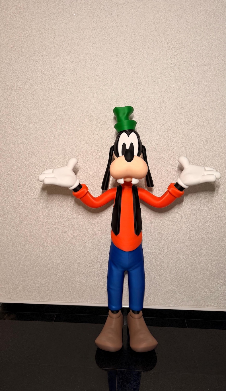 Puede incluir: Una figura de Goofy, un personaje de Disney, de pie con los brazos extendidos. La figura lleva un sombrero verde, orejas negras y un chaleco negro sobre una camisa naranja. Pantalones azules y zapatos marrones completan el look. La figura tiene guantes blancos y una gran sonrisa amistosa.