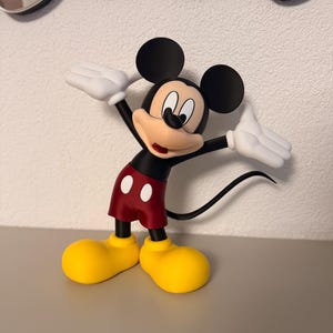 Puede incluir: Una figura de Mickey Mouse con los brazos extendidos. La figura presenta orejas negras, pantalones cortos rojos con botones blancos, zapatos amarillos y guantes blancos. La figura est&aacute; sobre una superficie gris.