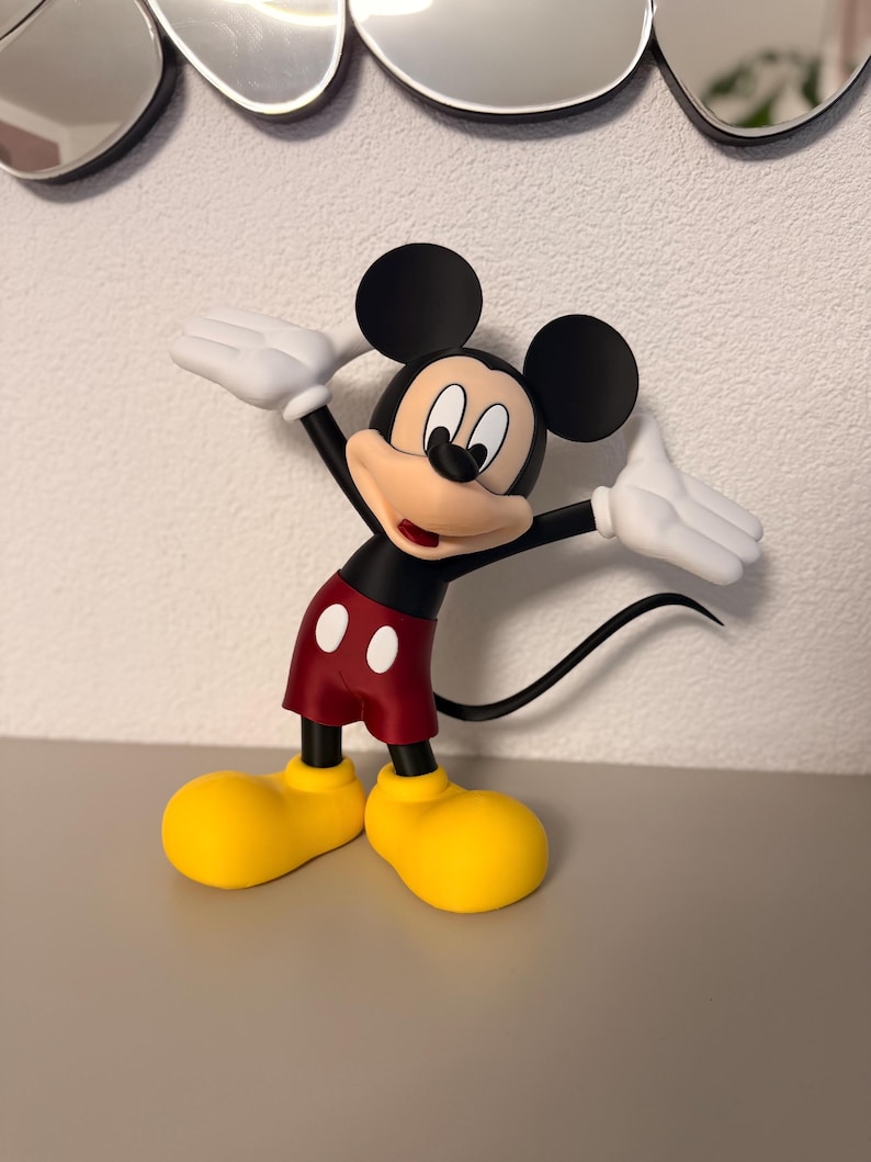 Puede incluir: Una figura de Mickey Mouse con los brazos extendidos. La figura presenta orejas negras, pantalones cortos rojos con botones blancos, zapatos amarillos y guantes blancos. La figura est&aacute; sobre una superficie gris.