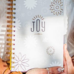 May include: A white spiral bound journal with a gold floral design and the word "JOY" in black lettering. The journal says "J-CURNAL" on the bottom.