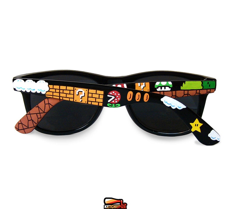 Picture of black sunglasses, hand-painted with a Super Mario 8 bit video game inspired design. Including 1up mushrooms, Piranha plant, Question blocks, Starman and Brick Blocks on a black background.
