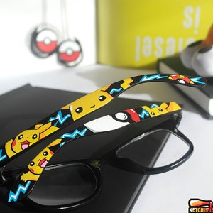 Picture of black clear lens glasses, hand-painted with a Pokemon design, including yellow Pikachu, blue and white bolts and red and white Pokeballs on a black background, in white, black, yellow, red and blue colors.