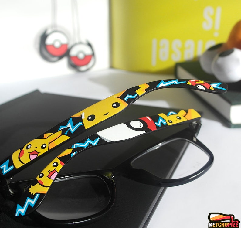 Picture of black clear lens glasses, hand-painted with a Pokemon design, including yellow Pikachu, blue and white bolts and red and white Pokeballs on a black background, in white, black, yellow, red and blue colors.