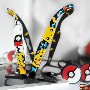 Picture of two black clear lens glasses, hand-painted with a Pokemon design, including yellow Pikachu, blue and white bolts and red and white Pokeballs, in white, black, yellow, red and blue colors.