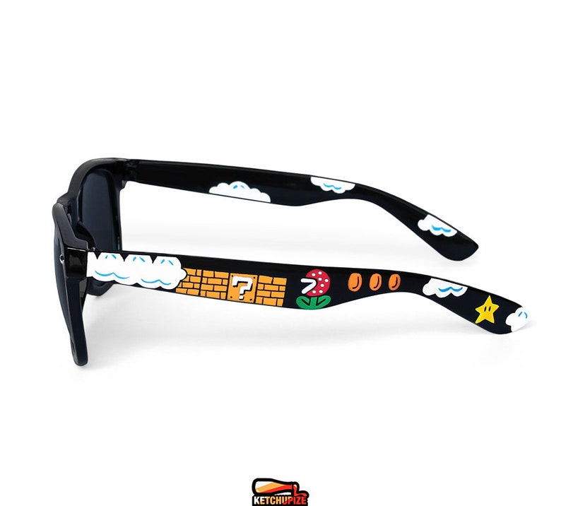 Picture of black sunglasses, hand-painted with a Super Mario 8 bit video game inspired design. Including 1up mushrooms, Piranha plant, Question blocks, Starman and Brick Blocks on a black background.