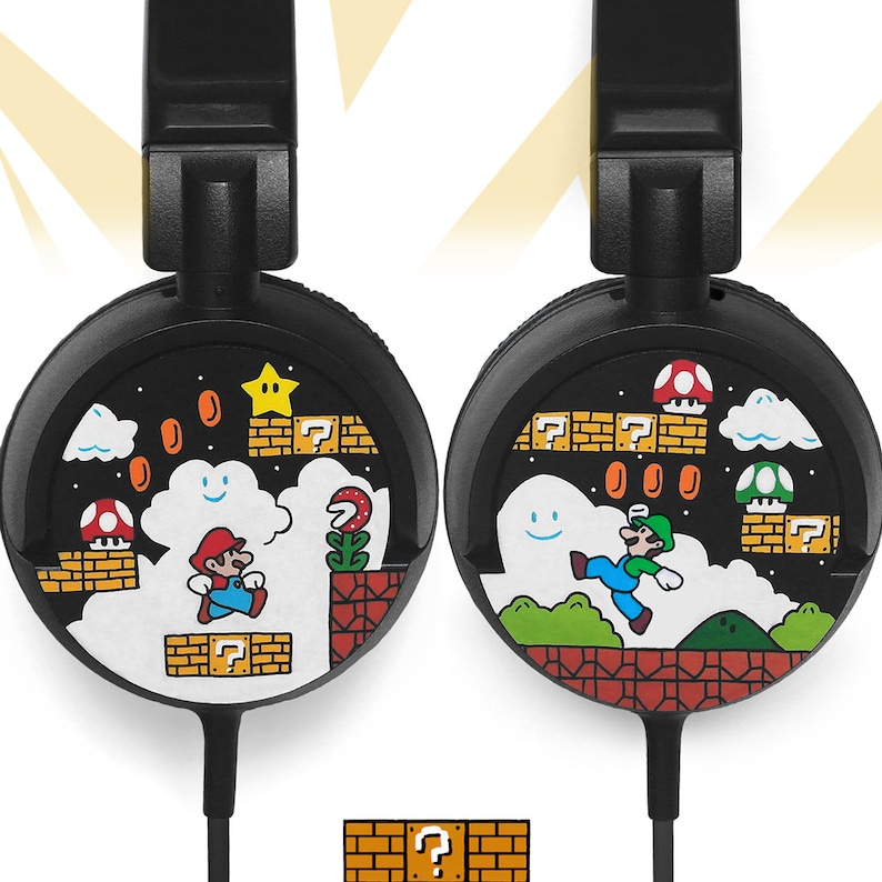 Picture of black headphones, hand-painted with a Super Mario 8 bit video game inspired design. Including the Mario brothers, Mario and Luigi, 1up mushrooms, Piranha plant, Question blocks, Starman and Brick Blocks on a black background.